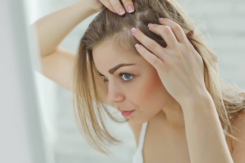A woman examines her scalp in the mirror, parting her hair with her hands. She appears focused, showcasing concern or curiosity. Soft lighting, neutral tones.