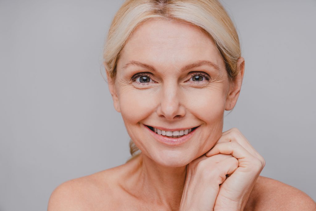 Smiling woman with light blonde hair against a soft gray background. She has a natural look and rests her chin gently on her hands, conveying warmth.
