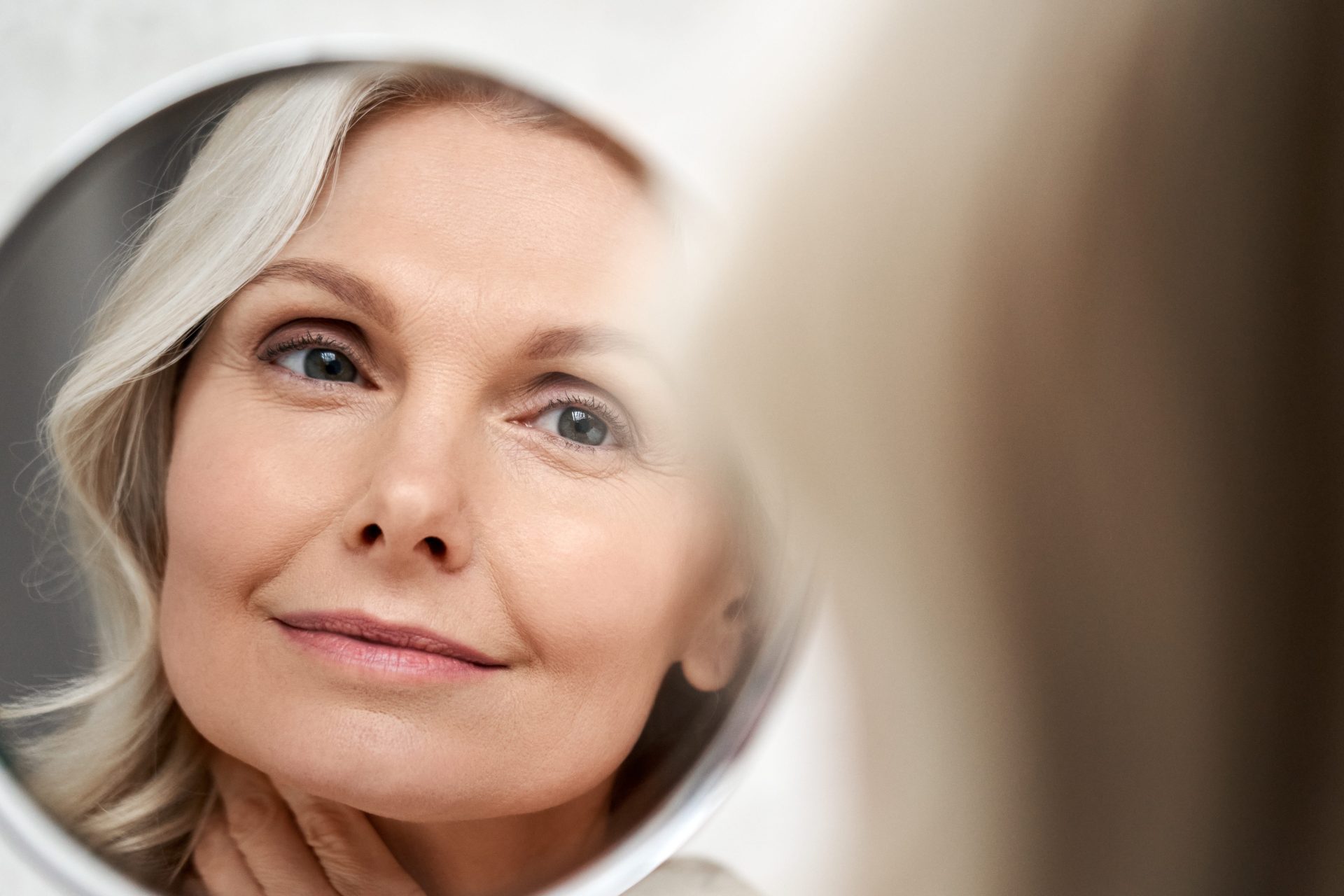 A woman with short, blonde hair looks at her reflection in a round mirror. She has a serene expression, conveying confidence and self-assurance.